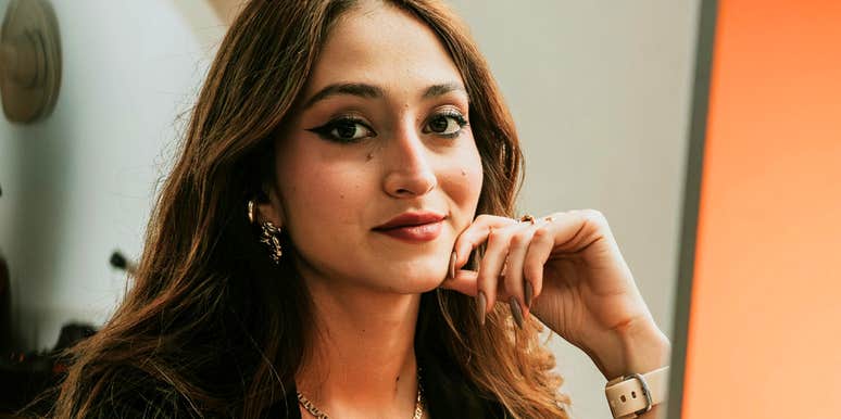 A woman sits indoors with her hand resting on her chin, looking toward the camera.