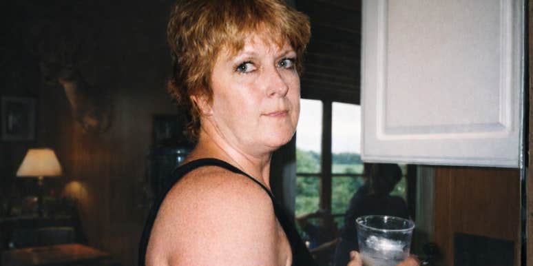 A woman standing indoors holding a glass, looking toward the camera.