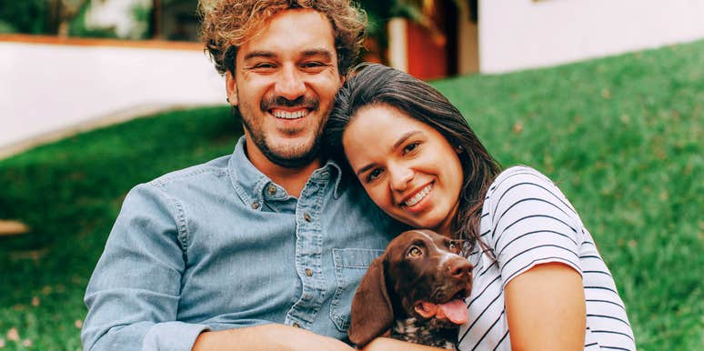 A smiling couple sitting on grass holding a small dog outdoors.