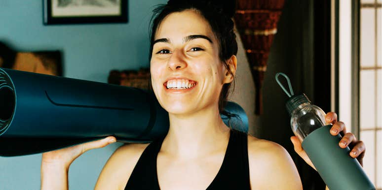 A woman smiling indoors holding a yoga mat and reusable water bottle.