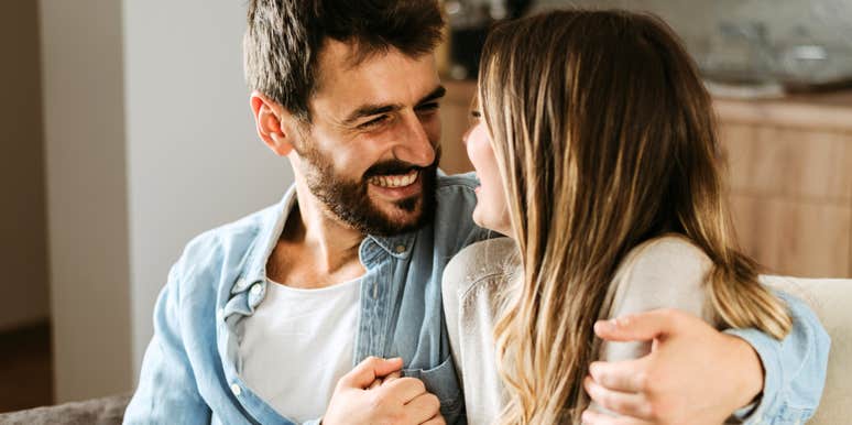 Man smiling warmly at a woman while sitting close together on a couch, sharing a relaxed, affectionate moment