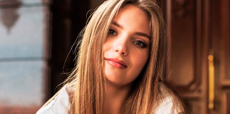 A woman with long hair looking calmly at the camera in a softly lit indoor setting.