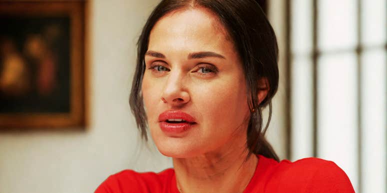 A woman with dark hair wearing a red top looking toward the camera indoors.