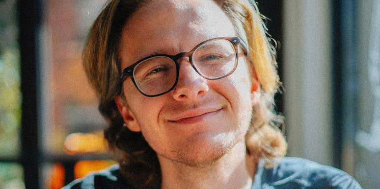 A person wearing glasses smiles calmly while sitting near a window in soft natural light.