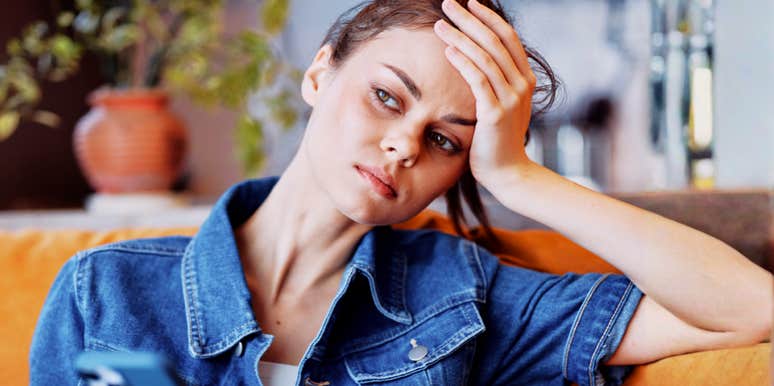 Woman in a denim shirt sitting on a couch, resting her head in her hand while looking at her phone with a worried, distant expression.
