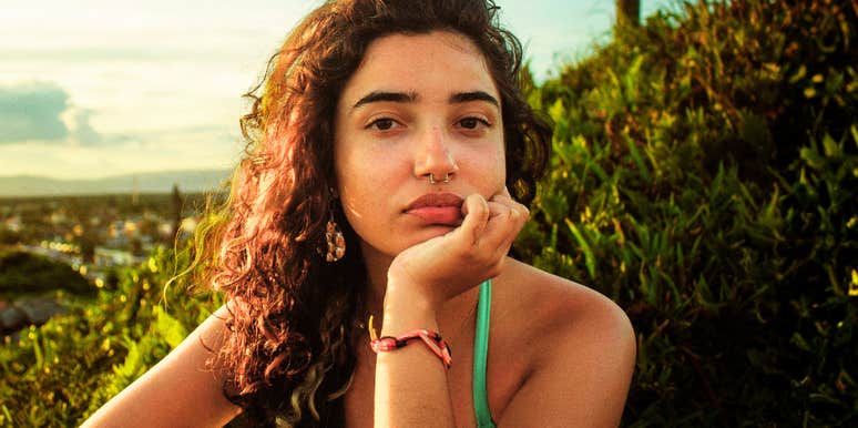 pensive young woman resting her chin on her hand at golden hour, gazing forward with distant, reflective expression against lush green backdrop