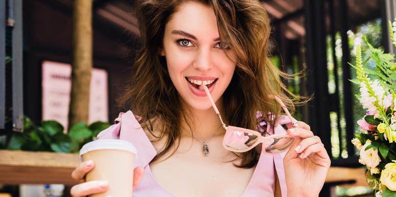 confident woman at outdoor café holding coffee and playfully biting sunglasses, flirty eye contact and relaxed body language capturing effortless charm and instant chemistry without small talk