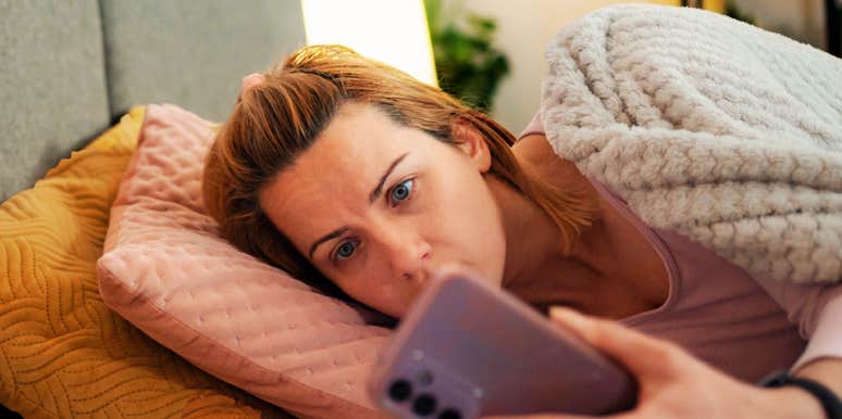 tired woman lying in bed under a blanket at night staring at her phone screen with wide, anxious eyes.