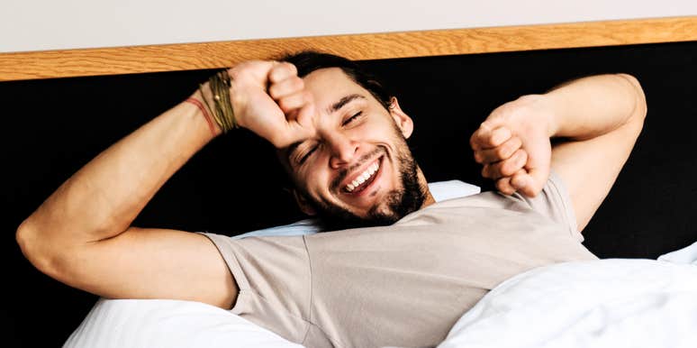 A man waking up in bed and stretching with a smile.