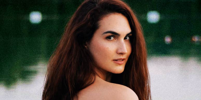 A close-up portrait of a woman with long brown hair looking over her shoulder outdoors.