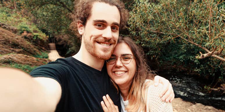 A couple taking a selfie outdoors near a wooded area.