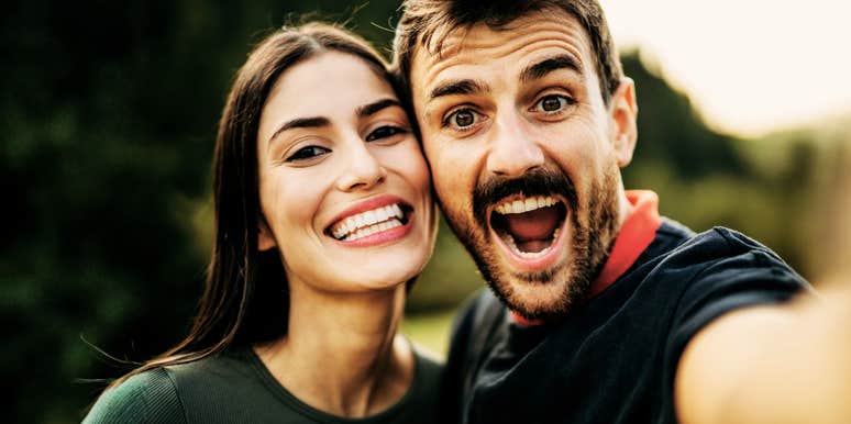 smiling couple taking close-up outdoor selfie in warm natural light, joyful authentic expressions, man with open-mouthed laugh and woman leaning in with bright smile.