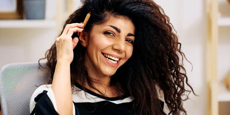 creative woman with curly hair smiling while holding a pencil near her temple, candid home office portrait capturing thoughtful expression and subtle habits associated with writers