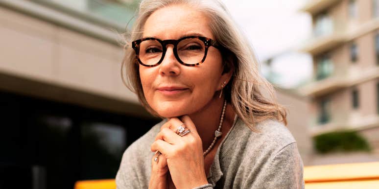 Senior woman with glasses sitting thoughtfully on a wooden bench in a city setting under a cloudy sky, reflecting on small life changes people make in their fifties and sixties to leave constant stress behind