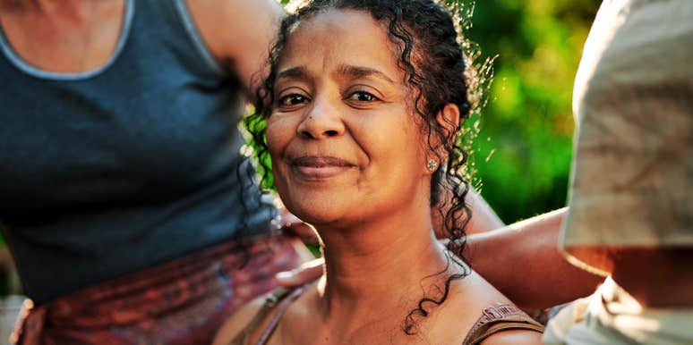A mature woman with a warm, radiant smile sitting outdoors at a wellness retreat, embodying the open and welcoming body language that instantly puts others at ease.