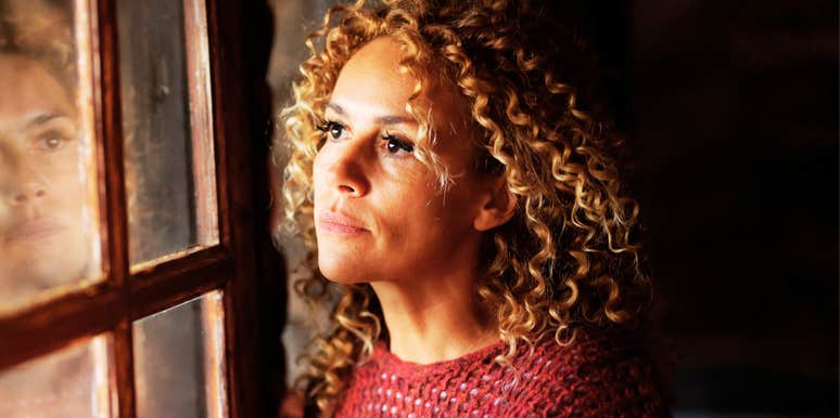 A thoughtful adult woman looking out a window with her reflection visible in the glass, illustrating the 'internalized habits' and subtle daily choices that lead to chronic unhappiness.