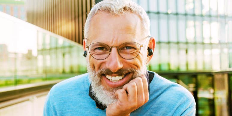 A mature man with a grey beard laughing and wearing headphones on a city street, illustrating the 'emotional vitality' and life satisfaction that peaks after fifty.