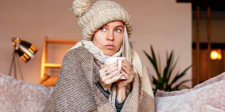 woman bundled up in hat and blanket holding mug sitting on couch at home