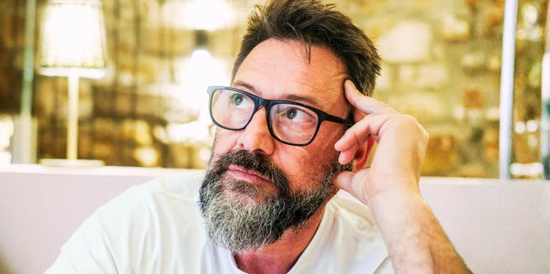 In a quiet room at home, a 50-year-old man with glasses sits alone, his face marked by sadness and concern, as he struggles with feelings of loneliness, emotional exhaustion, and personal worries
