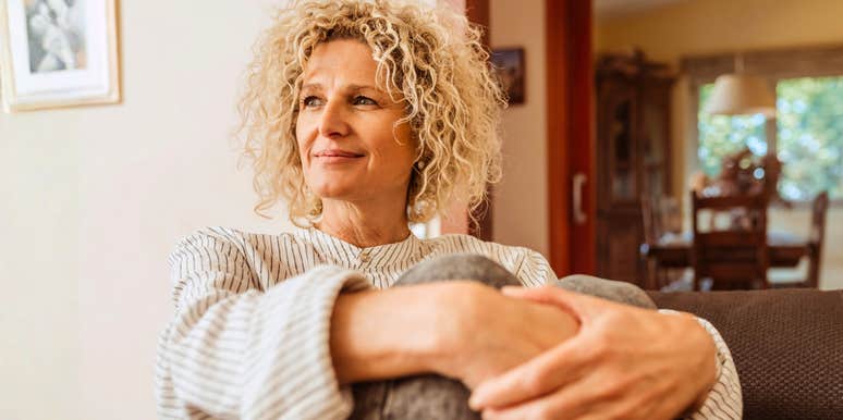 A stylish woman in her 50s relaxing in a modern living room with a warm, open expression; illustrating the 'community-first' approach to mental health that prioritizes belonging and shared experience.