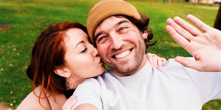 A joyful couple enjoying a day in nature, with the woman kissing the man’s cheek during a selfie; illustrating the spontaneous and consistent ways partners express their devotion and love every day.