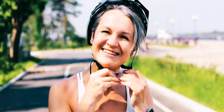Cheerful mature woman in a sports outfit fastening her bike helmet on a sunny day outdoors. She is standing on a paved cycling path, ready for a ride, showing an active and happy lifestyle.