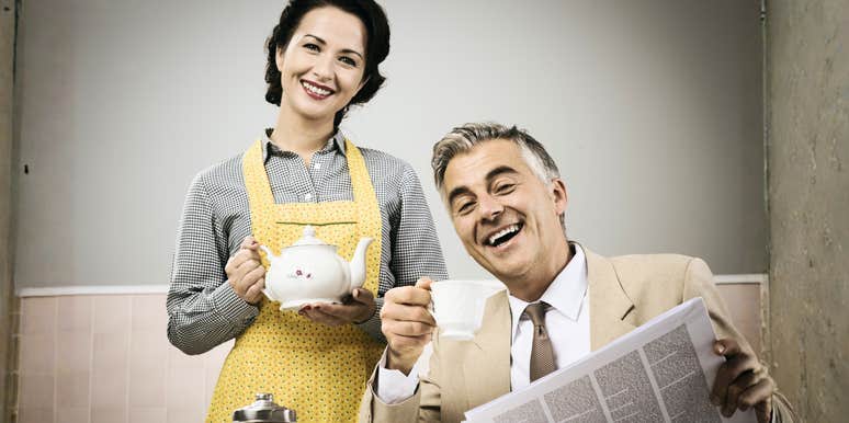 A staged vintage-style photo of a smiling woman pouring tea for her husband during breakfast; illustrating the rigid and often absurd domestic standards taught to women in the 1950s.