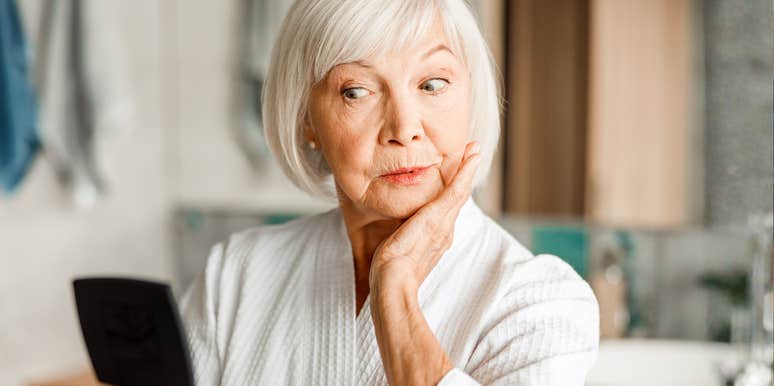 A portrait of a mature woman in a bathrobe preparing for a makeover in front of a mirror; illustrating the viral 'Glam-Ma' trend where eighty-year-olds reclaim their style and confidence through high-glamour transformations.
