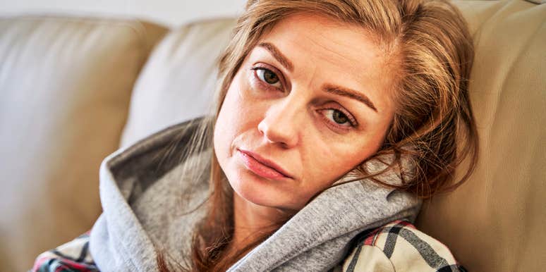 A woman sitting in solitude on a living room couch, looking depleted and overwhelmed; illustrating 'maternal burnout' and the systemic pressure on women to perform domestic happiness while struggling with internal depression.