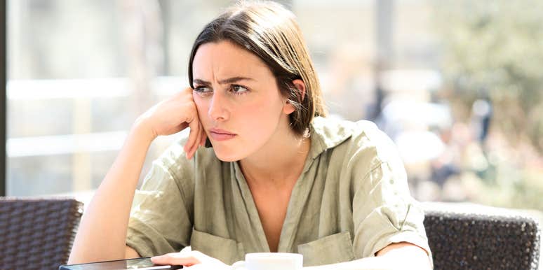 A woman looking frustrated and pensive while holding a smartphone in a social setting; illustrating 'reactive aggression' and the normalization of checking a partner's private messages or location out of suspicion.