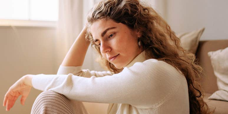 A woman sitting in a living room looking away with a sense of isolation; illustrating the 'silent erosion' of a relationship where communication has shifted from active engagement to habitual defensiveness and emotional distance.