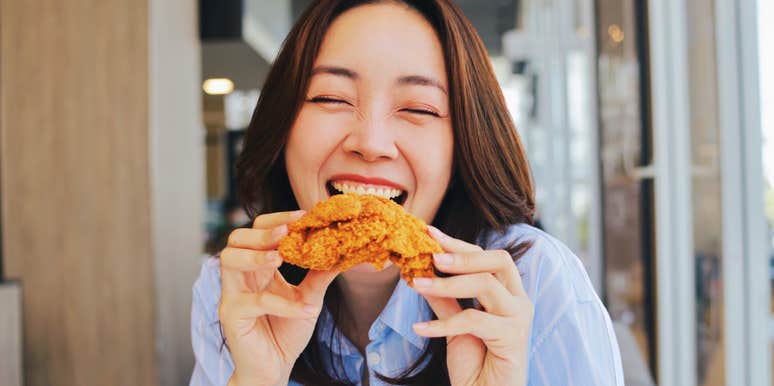 A joyful young Asian woman laughing wholeheartedly while holding a piece of crispy fried chicken at a restaurant; a visual representation of 'the marketed euphoria' contrasting with America's declining fast food quality.