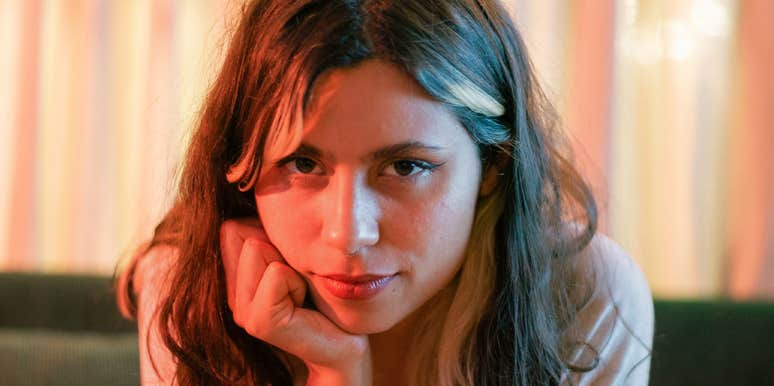 A woman sitting on a couch leaning forward with her hand on her chin, holding direct eye contact with a slightly sinister grin.