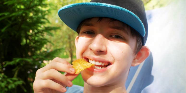 close-up lifestyle shot of a happy pre-teen boy eating snacks outdoors in a green space; illustrating 'the free-range autonomy deficit' where standard, unscripted childhood leisure is replaced by micro-managed scheduling.