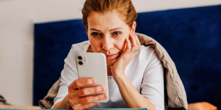 A woman lying in bed pulled under a heavy blanket, focusing intently on her glowing smartphone screen; a visual representation of 'the cloistered consumer' and the reality of millennial screen addiction.