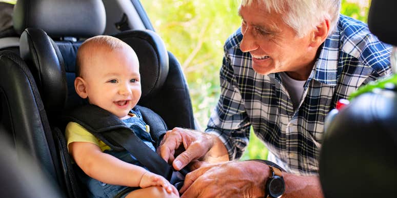 A happy senior man securing a cute toddler boy into a child safety seat for a road trip; illustrating a critical 'parental authority breach' and the breakdown of trust between parents and grandparents.