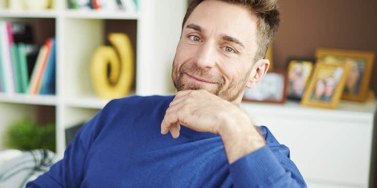 A gentle, smiling man sitting on a couch in a brightly lit room, capturing the 'psychological safety' and baseline warmth found in thriving, long-term relationships.