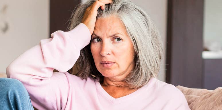 A pensive middle-aged woman in a cardigan rests her head on her hand while sitting on a living room couch; illustrating 'the midlife identity metamorphosis' where deep psychological restructuring occurs.