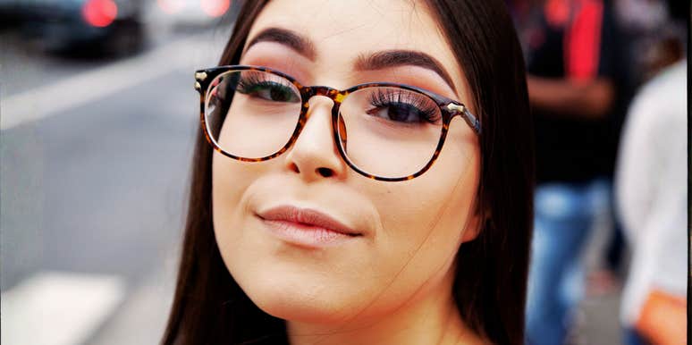 A close-up portrait of a thoughtful young woman wearing tortoiseshell glasses, a blue top, and a light trench coat with a patterned scarf, looking at camera with an observant, analytical expression on a city street.