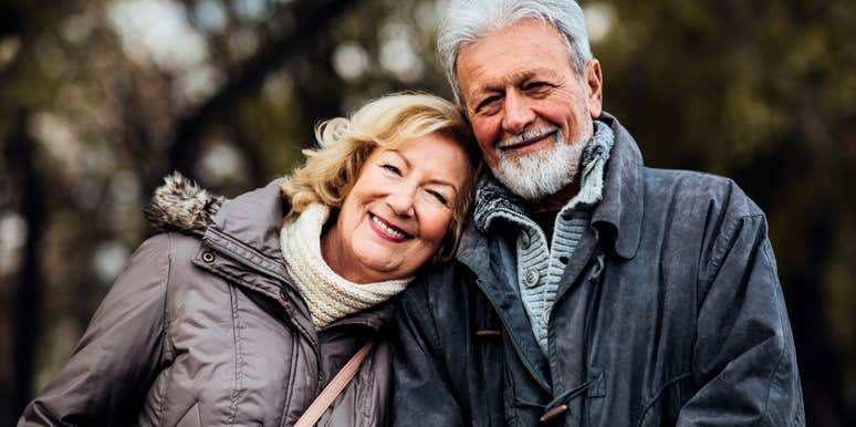 A happy married couple dressed in dark winter coats holding hands and walking along a wooded trail on a crisp day; a visual representation of the 'companions-in-motion' archetype used to study long-term relationship health.