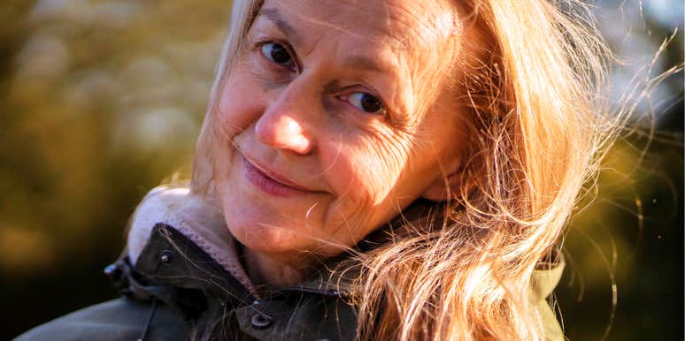 A close-up portrait of a beautiful mature woman with silver-streaked hair smiling serenely outdoors in golden autumn sunlight; a visual representation of 'the grounded elder' archetype used to study aging with dignity.