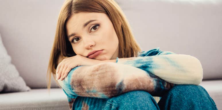 A young woman sitting in a cozy living room, leaning forward to rest her head on her folded arms over the back of a sofa, looking thoughtfully forward; a visual representation of 'the solitary reflector' archetype used to study emotional withdrawal in relationships.