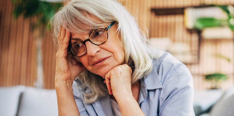 A somber senior woman sitting alone near a window at home, looking out pensively, conveying emotional isolation, loneliness, and reflection.