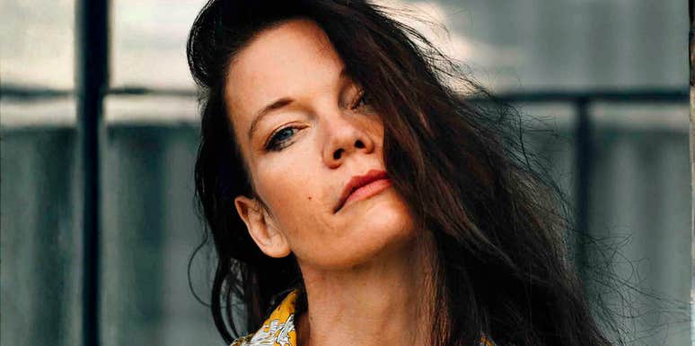 A candid outdoor street portrait of a fashionable young woman wearing a colorful printed top, looking directly into the camera with an intriguing, calm expression.