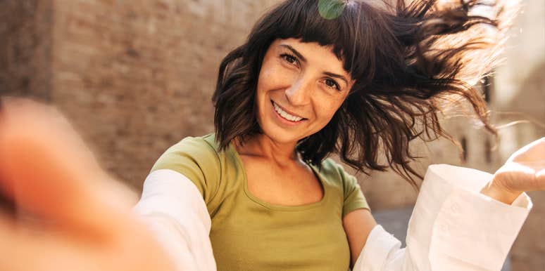 An outdoor portrait of a brunette woman with a bright, charming smile looking at the camera, reflecting high emotional intelligence, warmth, and interpersonal appeal.