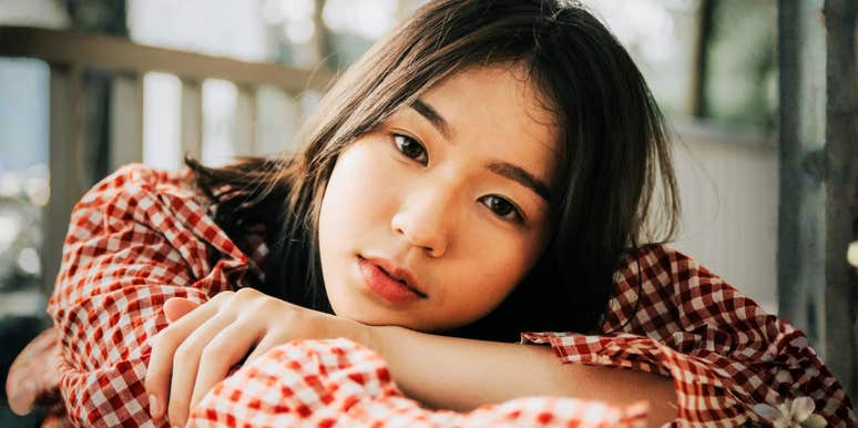 A close-up portrait of a young East Asian woman with straight dark hair wearing a red checkered shirt, leaning her head on her arms outdoors.