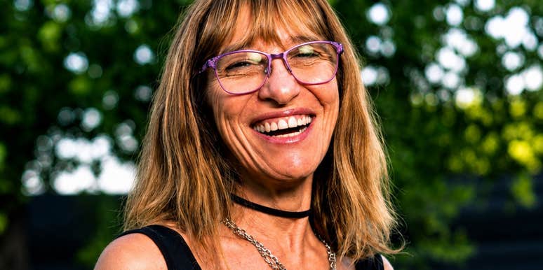 A woman with a choker necklace, chain, and purple eyeglass frames smiling broadly against lush green foliage, symbolizing humor, nostalgia, and generational perspective.