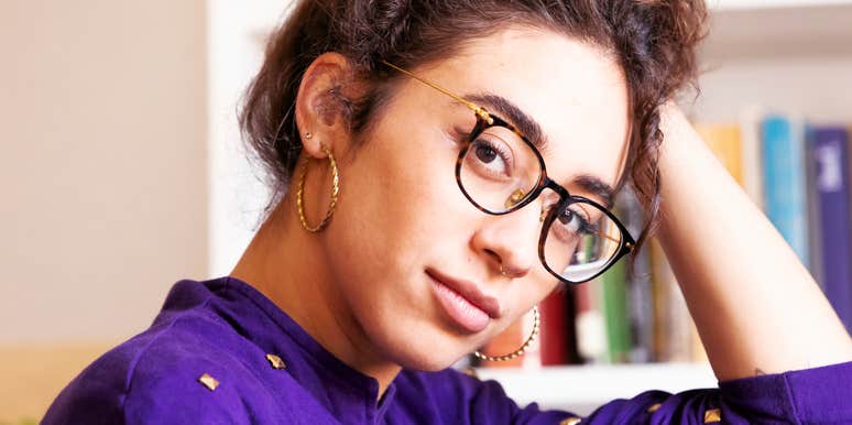 A close-up portrait of a woman with curly hair, tortoise-shell glasses, and gold hoop earrings, resting her hand behind her head while looking directly at the camera.