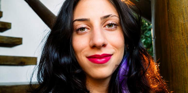 A woman with dark wavy hair and a calm, friendly expression looking into the camera outdoors near a wooden staircase, symbolizing practical logic, clarity, and ease.