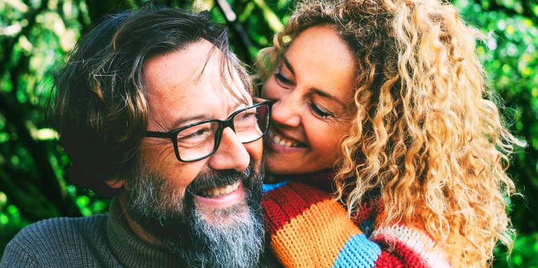 Smiling middle-aged couple laughing together outdoors, the woman leaning her cheek against the man’s, surrounded by green foliage.